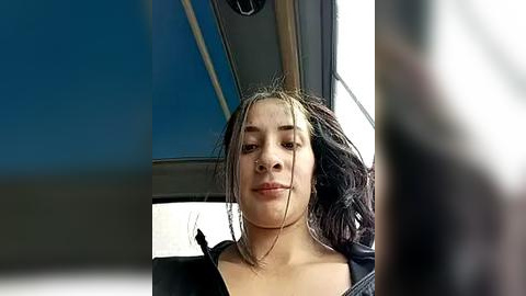 Media: Video of a young woman with wet, dark hair, wearing a black jacket, sitting in a car with a blurry, out-of-focus background.