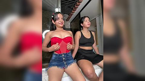 Media: Video of two women in modern, casual outfits: one in a red strapless top, denim shorts, and the other in a black crop top, high-waisted skirt, both with medium builds, sitting indoors.