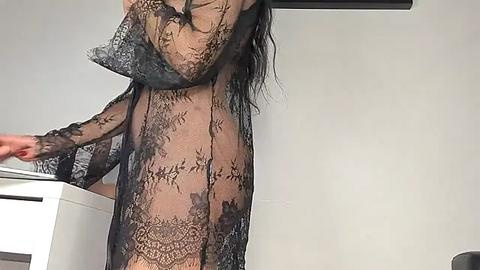 Media: Video of a slender, nude woman with long, dark hair, wearing a sheer black lace robe, adjusting her bra in a minimalist, modern room.