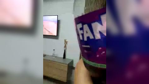 Media: A video shows a blurred person wearing a blue and white \"FAN\" hat in a modern room with a TV displaying a pink and purple screen, a wooden table, and a vase.