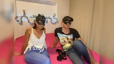 Media: Video of a woman and man in a room, wearing masks and casual clothes; pink bed, festive decorations, and a video game controller.