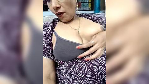 Media: Video of a Latina woman with light skin, wearing a black and white zebra print dress, lifting her left breast, revealing a grey bra. Background shows blurred objects and a blue wall.