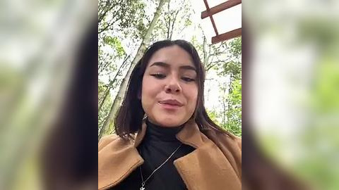 Media: Video of a Latina woman with medium skin tone, straight dark hair, wearing a brown coat and black turtleneck, smiling confidently. Background features lush green foliage and a wooden structure, suggesting an outdoor setting.