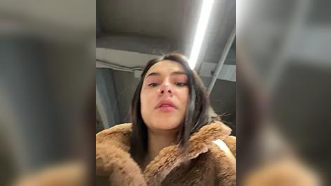 Media: Video of a young woman with long, dark hair, wearing a brown fur coat, looking down. She has a neutral expression. The background is a dimly lit, modern indoor space with metallic elements and fluorescent lighting.