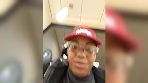 Media: A blurry video of a person wearing a red hat, glasses, and a black jacket, possibly in a recording studio with microphones and a reflective ceiling.