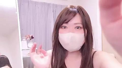 Media: Video of an East Asian woman with long brown hair, wearing a white face mask, brown top, and making a hand gesture in a brightly lit room with white walls and a gray curtain.