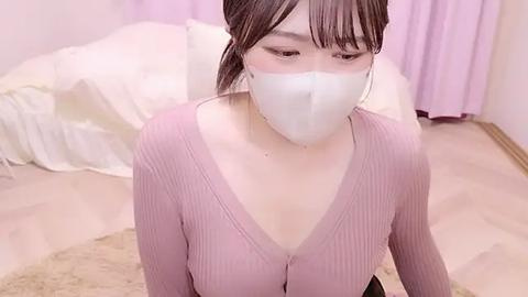 Media: Video of an Asian woman with light skin and brown hair in a ponytail, wearing a white face mask, a pink ribbed long-sleeve top, and a beige carpet in a softly lit, pastel-colored bedroom with white sheets.