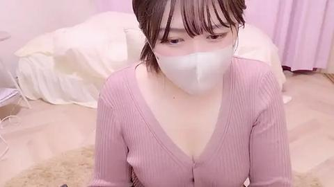 Media: Video of an East Asian woman with pale skin, wearing a pink ribbed sweater, white face mask, and straight black hair, sitting on a beige carpet in a pink room with a white bed and curtains.