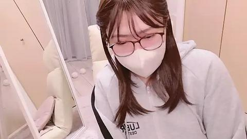 Media: Video of a young Asian woman with straight brown hair, wearing glasses and a white face mask, sitting in a white room with beige walls and a mirror, holding a pink object.