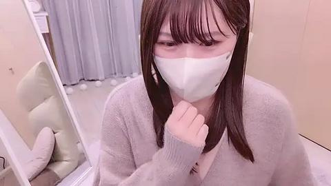 Media: A video of a young Asian woman with long black hair, wearing a light gray cardigan, white face mask, and a white T-shirt, sitting in a room with a beige carpet and light-colored curtains.