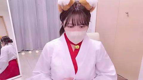 Media: Video of an Asian woman wearing a white lab coat, red blouse, and cat ears, with a mask, seated at a desk with a mirror.