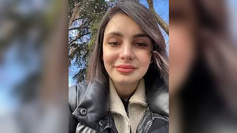 Media: A video of a young woman with light brown skin, straight dark brown hair, and light makeup, wearing a black leather jacket over a beige sweater, smiling in a forest with a clear blue sky.