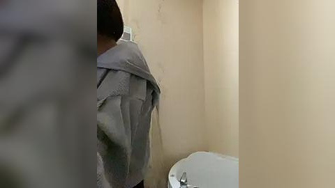 Media: Video of a man in a gray hoodie, back facing the camera, standing in a beige-walled bathroom with a white toilet visible on the right.