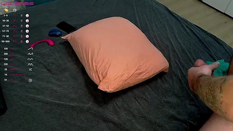 Media: A video of a man's hand, wearing a green glove, holding a pink pillow on a dark gray bed. A smartphone with a pink case and a black remote control are nearby.