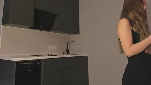 Media: Video of a woman in a black dress, standing in a modern kitchen with sleek black cabinets and a white countertop, featuring a black faucet.