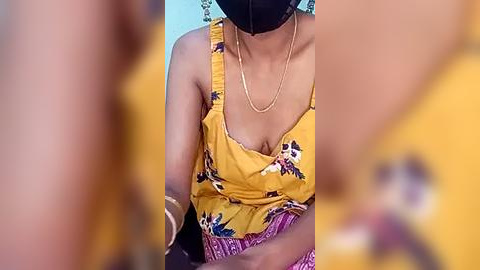 Media: Video of a woman with medium brown skin, wearing a yellow floral top, black mask, and gold necklace, seated in a yellow background, blurred hands in foreground.