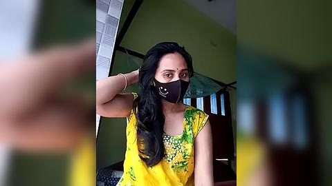 Media: Video of a woman with long black hair and fair skin wearing a yellow blouse with green floral patterns, black mask, and a yellow sari, posing indoors with blurred background.