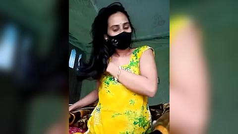 Media: Video of a woman with long black hair, wearing a yellow floral dress, black face mask, and black bracelets, adjusting her hair in a dimly lit room with green walls.