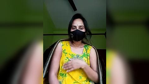 Media: A video shows a South Asian woman with medium skin tone, wearing a yellow floral blouse, black face mask, and black hair. She sits in a dark chair, hands clasped, in a dimly lit room with green walls.