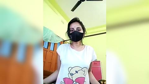 Media: Video of a woman wearing a black face mask, white t-shirt with cartoon character, in a bright, yellow-painted room with a fan and wooden furniture.