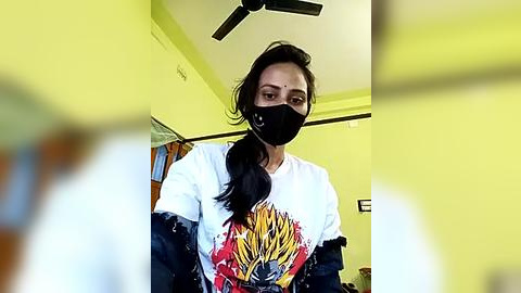 Media: Video of a young woman with medium skin tone, long dark hair, wearing a black face mask, white graphic tee, and dark blue jacket, indoors with a yellow wall and ceiling fan.