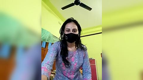 Media: A video of a woman in a mask, wearing a colorful, long-sleeved shirt, standing indoors with yellow walls, a fan, and furniture blurred in the background.