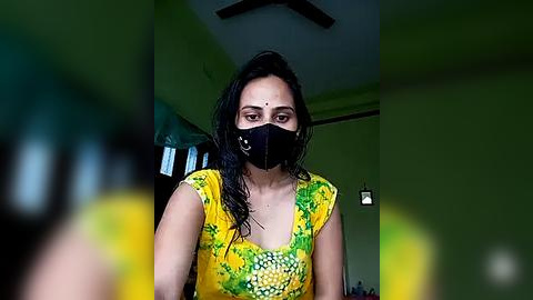 Media: Video of a South Asian woman with medium brown skin, wearing a black face mask, yellow floral dress, and black hair, in a dimly lit room with green walls.