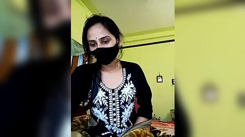 Media: Video of a woman in a black mask, black top with white embroidery, and a black headband, seated in a dimly-lit room with yellow walls and a bed with a brown blanket.