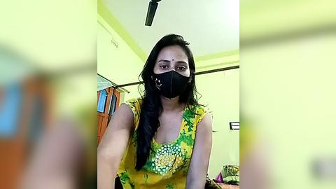 Media: Video of a South Asian woman with long black hair, wearing a yellow floral blouse, black mask, and forehead bindi, in a brightly lit room with green walls and wooden furniture.