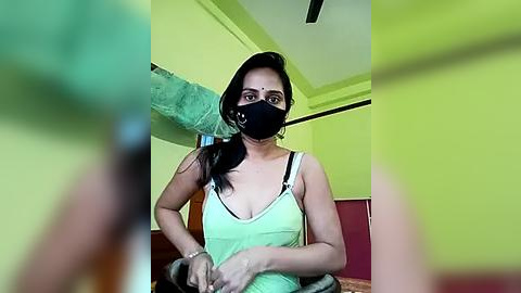 Media: A video of a woman with long black hair, wearing a black face mask, a green tank top, and a bracelet, seated indoors, with a blurred foreground and bright yellow walls.