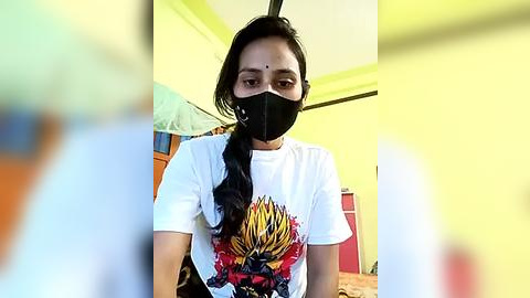 Media: Video of a South Asian woman with medium brown skin, long black hair, and a black face mask, wearing a white T-shirt with a colorful graphic design. She stands in a bright, yellow-walled room with a bed, red cabinet, and green curtains.