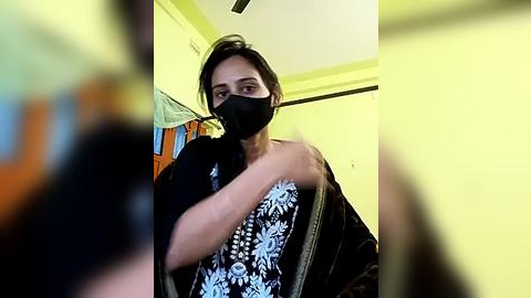 Media: Video of a young woman with medium skin tone, dark hair, wearing a black face mask and a black floral-patterned top, seated indoors against a yellow wall.