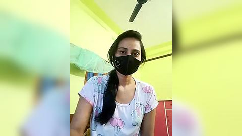 Media: Video of a woman with dark hair wearing a black face mask, white T-shirt with pastel balloons, and yellow walls. Background includes a fan and a partially visible bed with a green sheet.