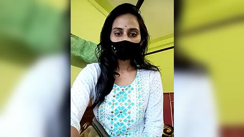 Media: Video of a South Asian woman with long black hair, wearing a white floral-patterned blouse, black mask, and yellow headscarf. Background features yellow walls and a blurred green plant.