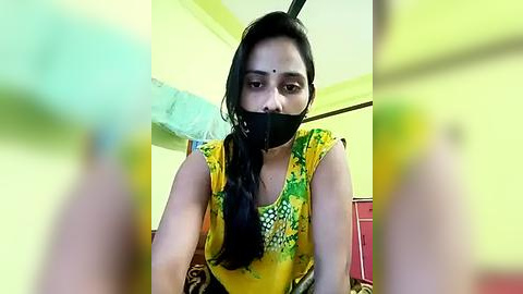 Media: Video of a South Asian woman with long black hair, wearing a yellow blouse with green patterns, black tape over her mouth, and a pink bindi, sitting on a bed in a yellow-painted room.