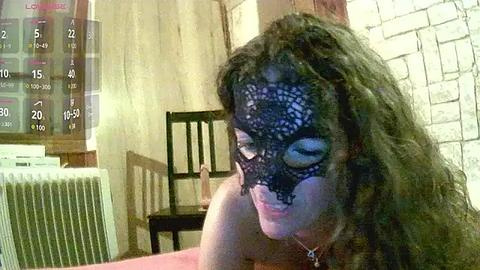 Media: A video of a woman with curly brown hair, wearing a black lace mask, sitting indoors next to a calendar, a radiator, and a wooden chair.