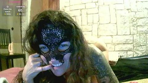 Media: Video of a woman with long, dark hair, wearing a black lace mask, lying on a bed with a stone wall backdrop, holding a phone camera, in a dimly lit room.