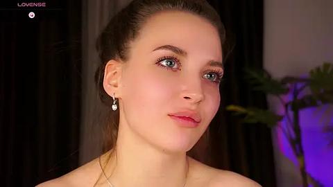 Media: Video of a fair-skinned, young woman with brown hair pulled back, wearing a strapless dress, earrings, and subtle makeup, indoors with a blurred background.