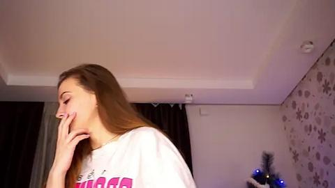 Media: Video of a young Caucasian woman with long brown hair, wearing a white t-shirt, covering her mouth with her hand, in a dimly lit room with floral wallpaper and dark curtains.