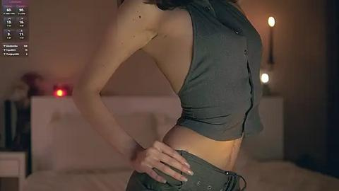 Media: Video of a slender, light-skinned woman in a sleeveless, button-up top, revealing her midriff, and wearing low-rise jeans. Background shows a dimly lit room with a bed and a lamp.