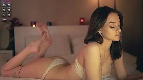 Media: Video of a slender, fair-skinned woman with long dark hair lying on her stomach in beige lingerie, feet playfully raised, in a dimly lit bedroom with a white bed and a wall-mounted lamp.