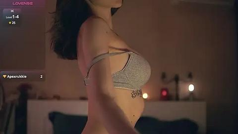 Media: A dimly-lit video of a pregnant woman in a grey bra, standing sideways in a bedroom with soft lighting, blurred background.