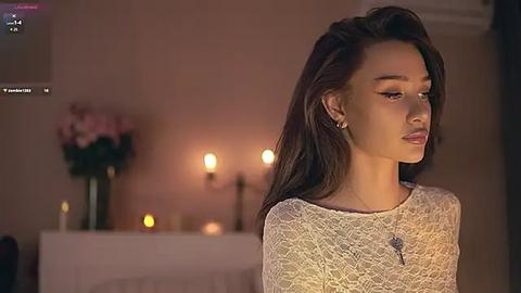 Media: Video of a young woman with long dark hair, wearing a lace top, in a dimly lit room with candles, a vase of pink flowers, and a cross on a shelf in the background.