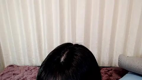 Media: A video of a person with long, straight black hair, sitting on a bed with a mauve quilted bedspread. The background features cream-colored vertical blinds. The person\u2019s back is to the camera.