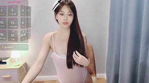 Media: Video of an East Asian woman with long, straight black hair, wearing a light pink, lacy bralette. She stands in a modern bedroom with a calendar and blue curtains in the background.