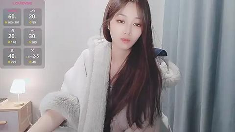 Media: Video of an East Asian woman with long, straight brown hair, wearing a white fur-trimmed coat, standing indoors beside a table lamp and gray curtain.