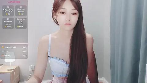 Media: Video of a young East Asian woman with long, straight black hair, wearing a light blue bralette and light pink shorts, standing in a modern, minimalist room with white walls and a blue curtain.