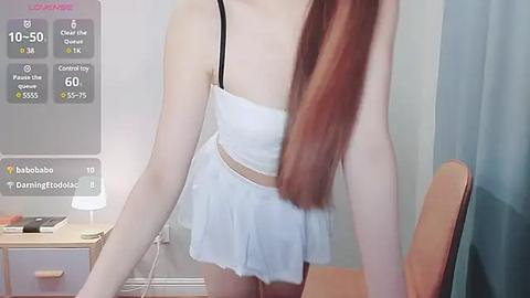 Media: A video of a young, slim Asian woman with long, straight red hair, wearing a white, spaghetti-strap dress, standing in a minimalist bedroom with a wooden chair and a lamp.