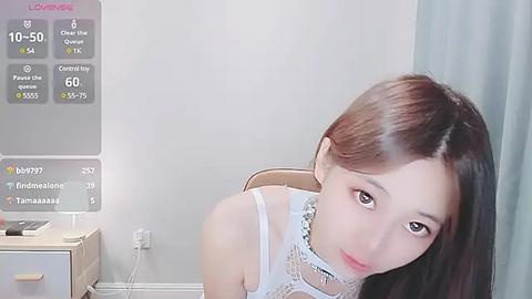 Media: Video of a young East Asian woman with long brown hair, wearing a white lace top, looking at a digital display with health stats, in a minimalist room with a light blue curtain.