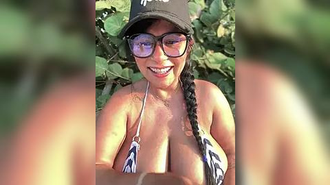 Media: Video of a smiling, tan-skinned woman with large breasts, wearing a black baseball cap and sunglasses, in a leafy outdoor setting.